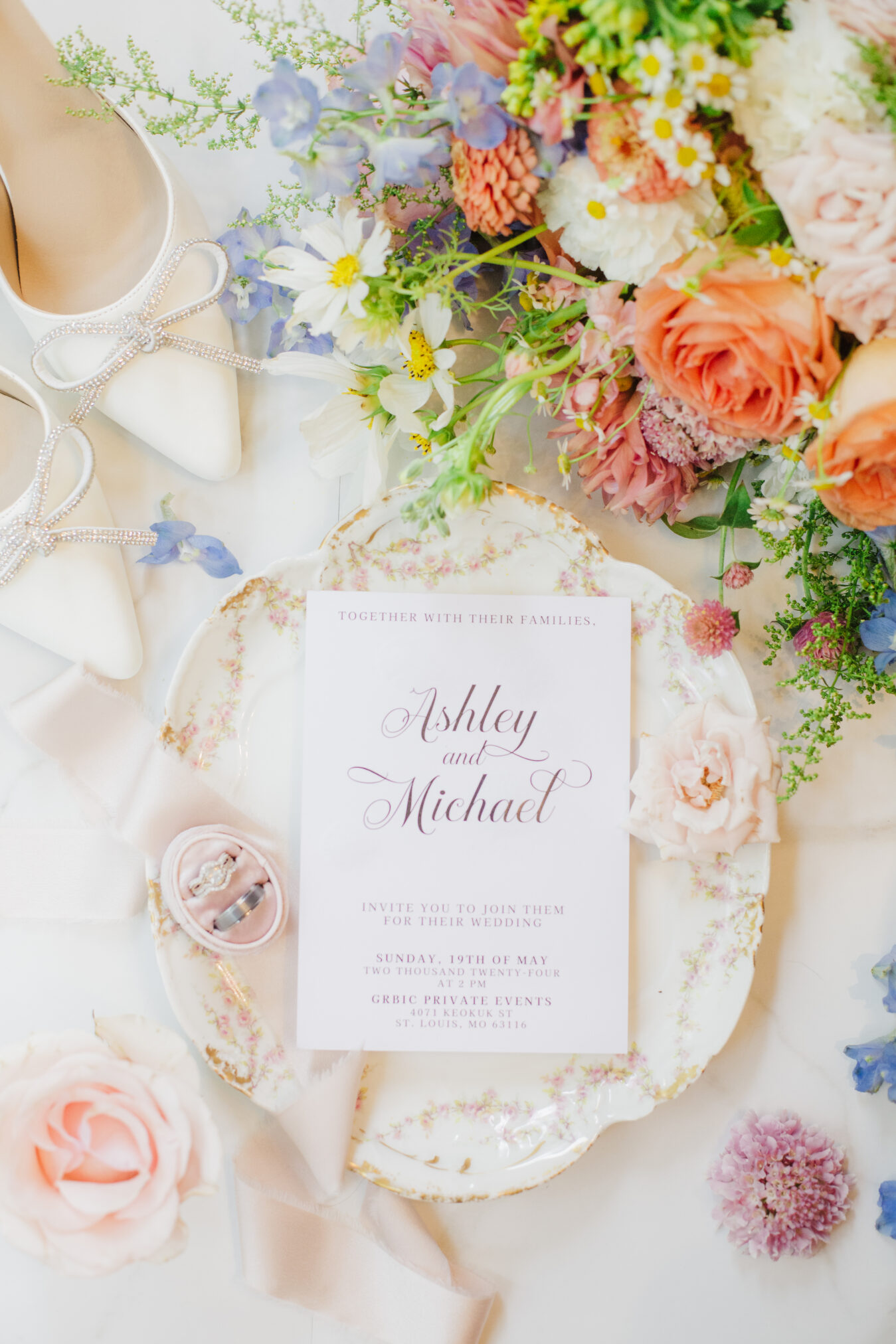 Flatlay with wedding invitation, flowers, high heels, and rings.