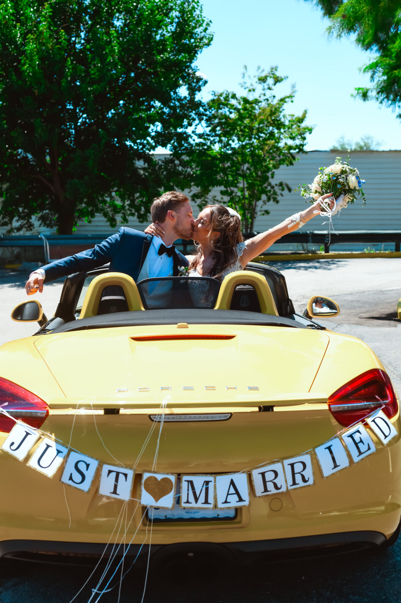 Bride holds bouquet with groom in a yellow convertible with a 'Just Married' sign attached to the car.